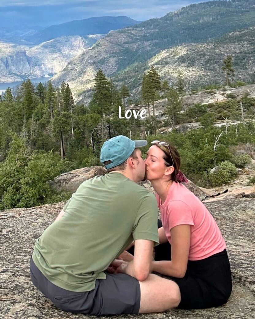 Man in swim trunks holds a baby in a blue life jacket in shallow water.