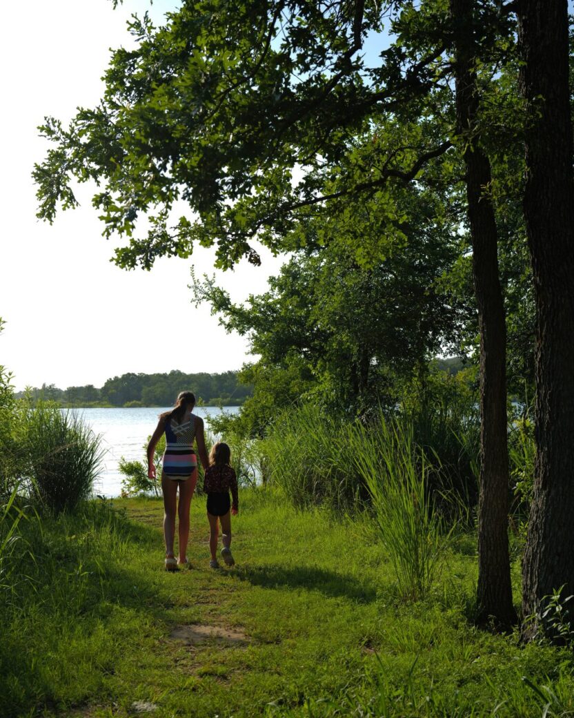 Man in swim trunks holds a baby in a blue life jacket in shallow water.