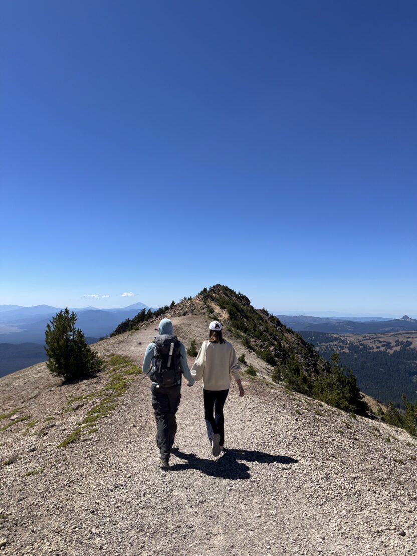 Man in swim trunks holds a baby in a blue life jacket in shallow water.