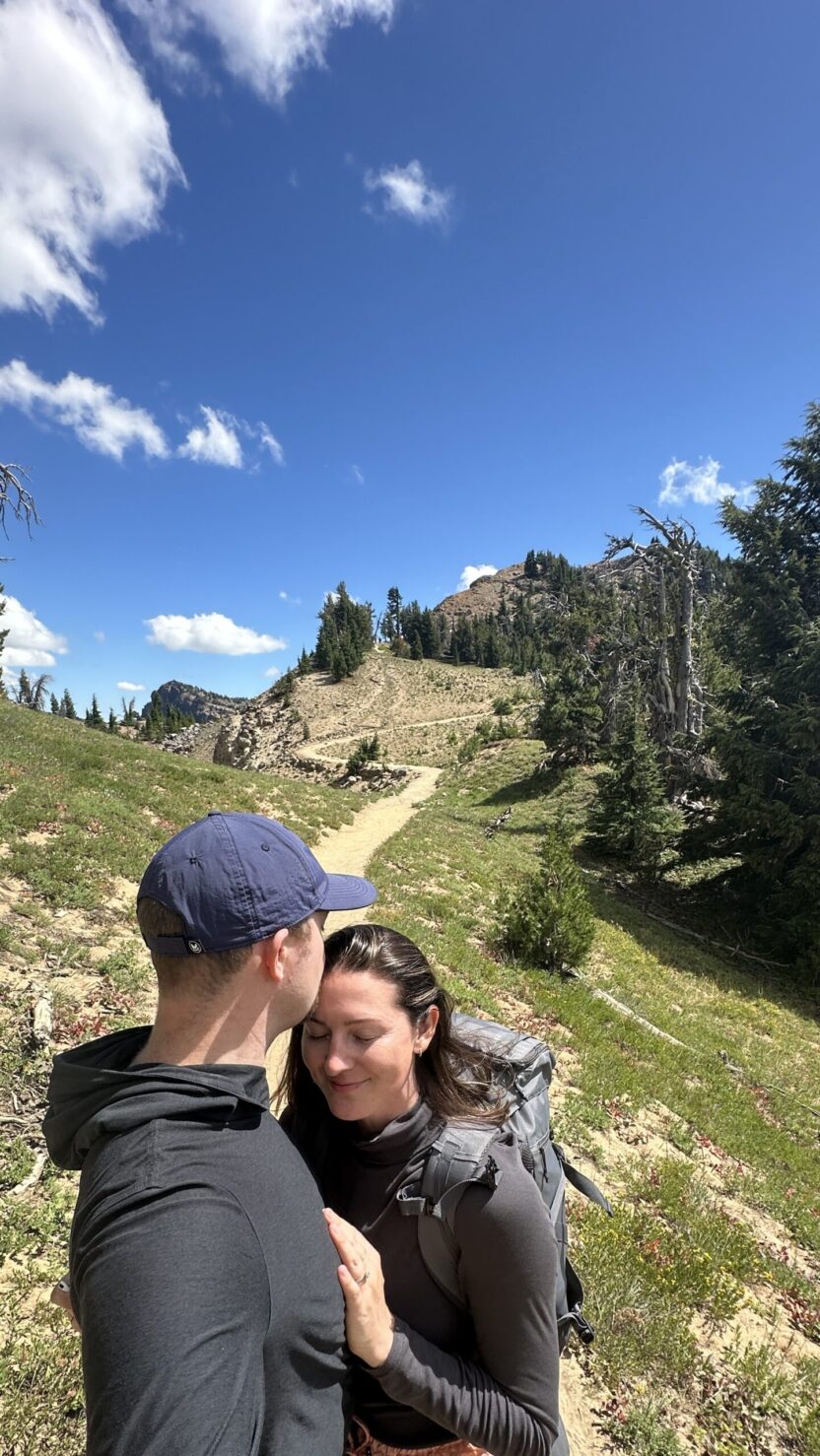 Man in swim trunks holds a baby in a blue life jacket in shallow water.