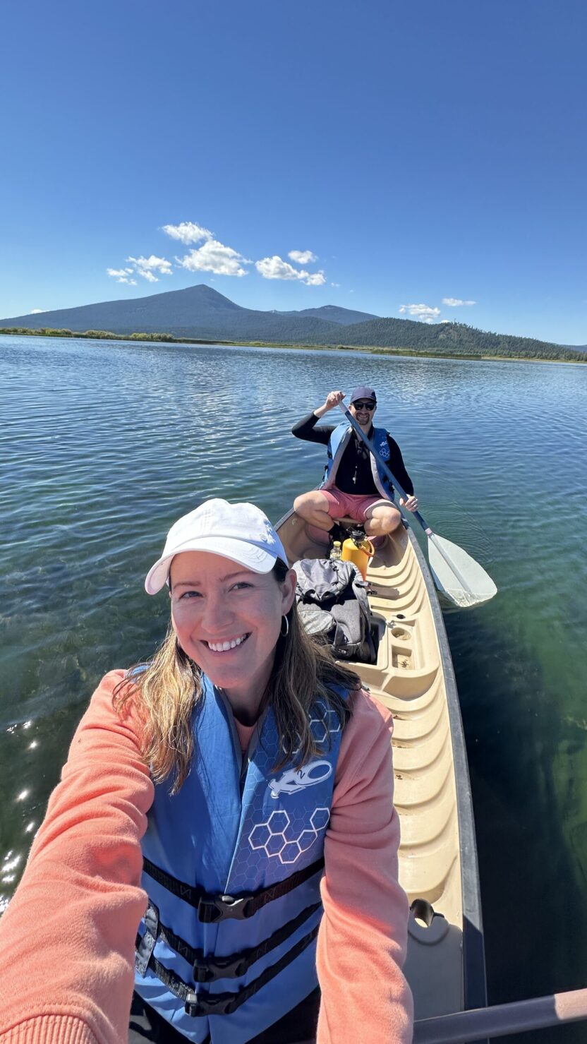 Man in swim trunks holds a baby in a blue life jacket in shallow water.
