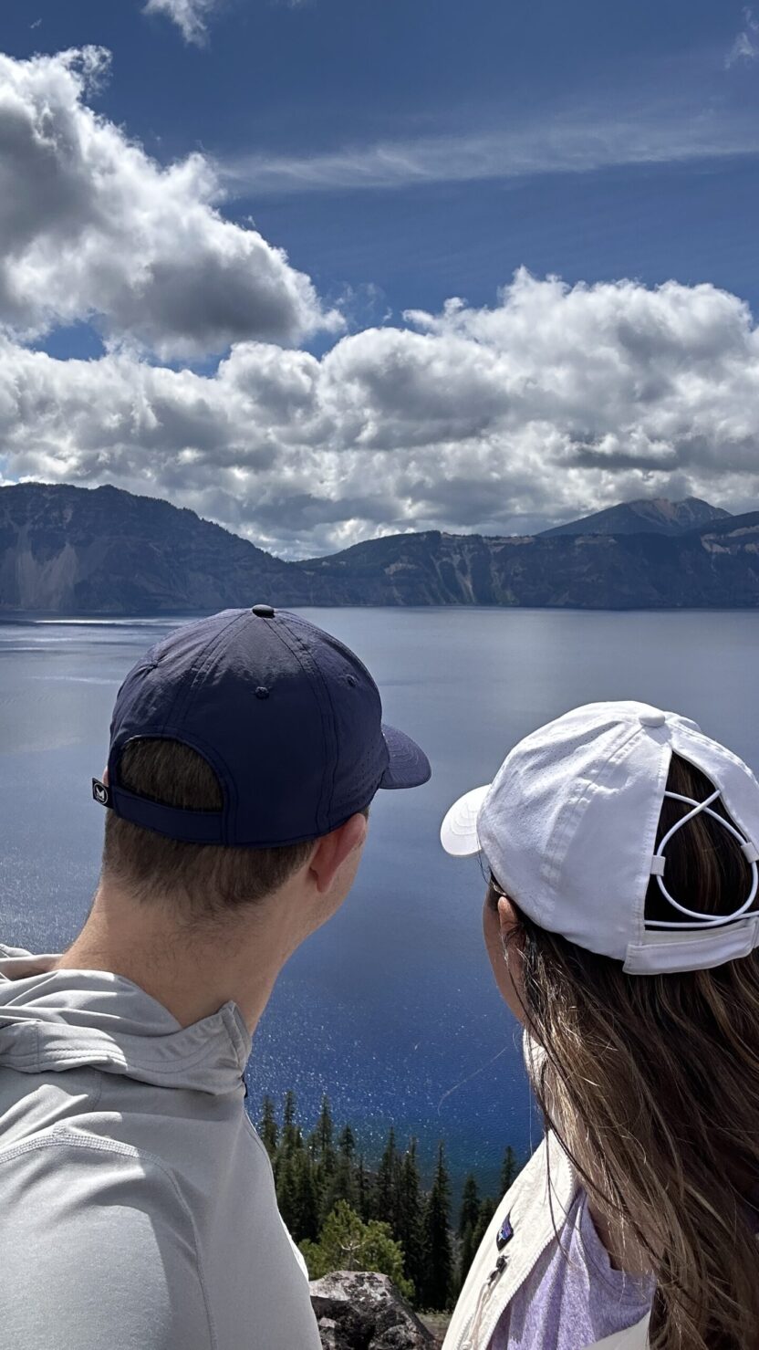 Man in swim trunks holds a baby in a blue life jacket in shallow water.