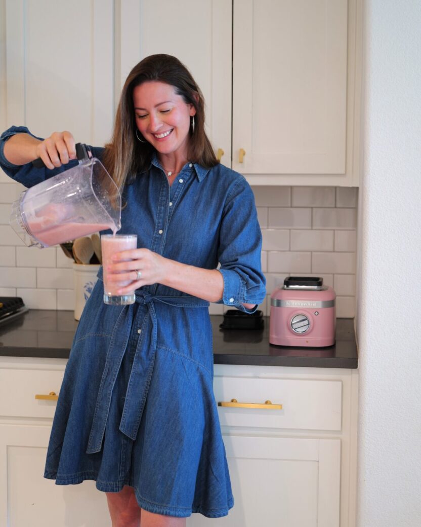 Man in swim trunks holds a baby in a blue life jacket in shallow water.
