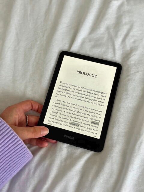 flat lay picture of a hand holding a kindle. Part of the purple sweater is showing. The book on the kindle is promoting the April 2026 Book of the month pick. It's called Theo of Golden.