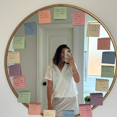 Chic girl smiling wearing white. Mirror selfie. Mental health affirmations and faith affirmations are written in cursive on muted rainbow colored sticky notes taped to a big, gold, round mirror.