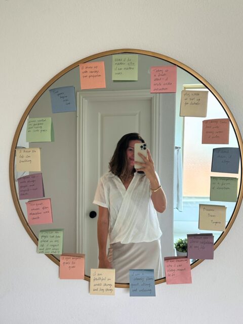 Chic girl smiling wearing white. Mirror selfie. Mental health affirmations and faith affirmations are written in cursive on muted rainbow colored sticky notes taped to a big, gold, round mirror.