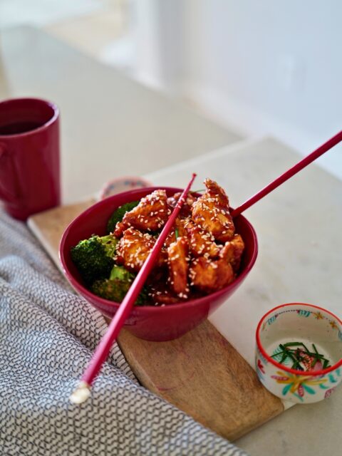 honey garlic chicken and broccoli in a red bowl with chopsticks on a kitchen counter